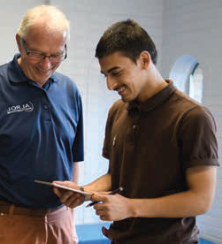 Cerbando Trejo smiling, holding a pen and ledger, showing his work to an older gentleman